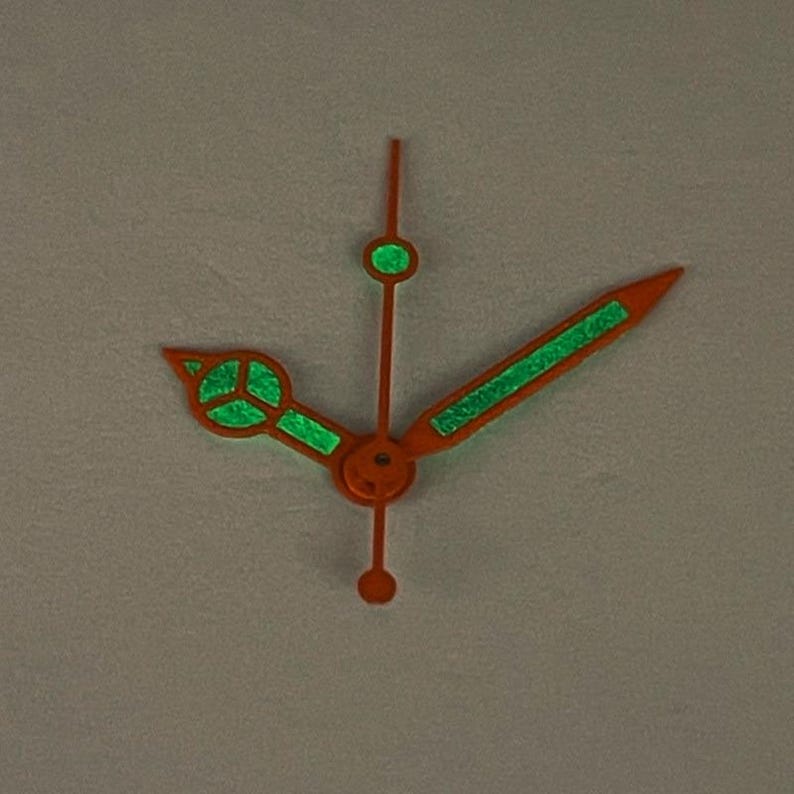 May include: Orange watch hands with glowing green accents. The hands are a vibrant orange color, with luminous green detailing. The design includes a second hand, minute hand, and hour hand, all with a unique, eye-catching style. The hands are designed for a watch.