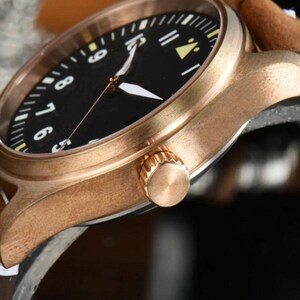May include: Close-up of a bronze-colored wristwatch with a black face and white numerals. The watch has a brown leather strap and a gold-colored crown. The watch hands are white.