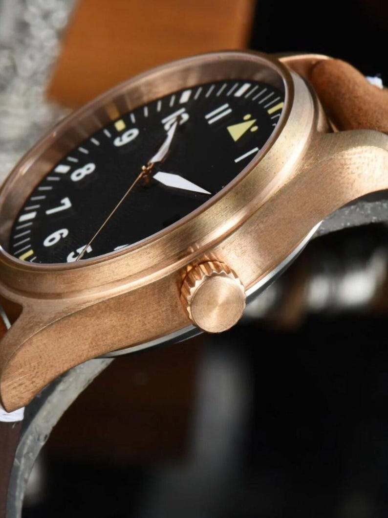 May include: Close-up of a bronze-colored wristwatch with a black face and white numerals. The watch has a brown leather strap and a gold-colored crown. The watch hands are white.