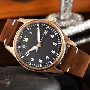 May include: A bronze-colored wristwatch with a black face and white numerals. The watch has a brown leather band with white stitching. The watch face displays the date and has luminous hands. The watch is a classic, timeless design.