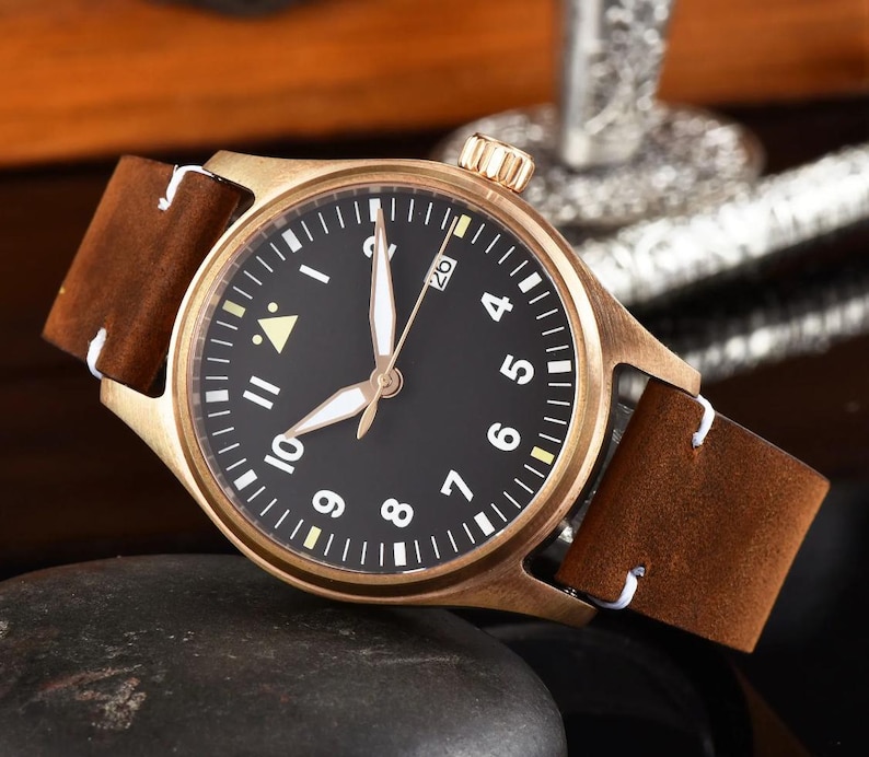 May include: A bronze-colored wristwatch with a black face and white numerals. The watch has a brown leather band with white stitching. The watch face displays the date and has luminous hands. The watch is a classic, timeless design.