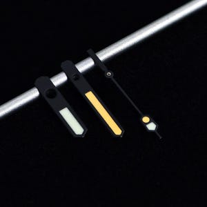 May include: Close-up of three black watch hands with luminous accents. The hands are in different styles, with white, yellow, and a combination of white and yellow. The hands are set against a dark background and a silver bar.