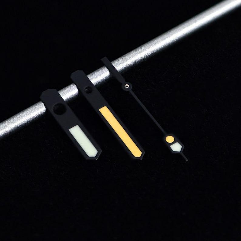 May include: Close-up of three black watch hands with luminous accents. The hands are in different styles, with white, yellow, and a combination of white and yellow. The hands are set against a dark background and a silver bar.