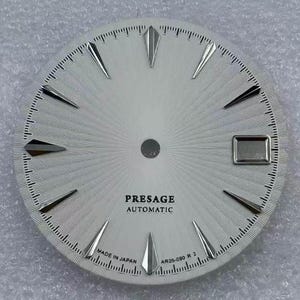 Könnte beinhalten: Ein rundes, silberweißes Zifferblatt mit Strahlenmuster, silberfarbenen, facettierten Stundenmarkierungen und einem rechteckigen Datumsfenster auf der Drei-Uhr-Position. Der Text PRESAGE AUTOMATIC, MADE IN JAPAN und AR05-090 R 2 ist schwarz gedruckt.