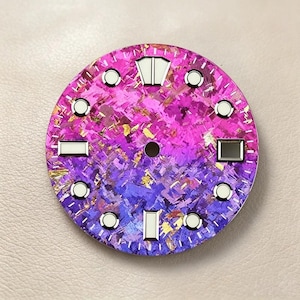Puede incluir: Una esfera de reloj circular con un degradado vibrante de rosa y morado, un acabado texturizado y fragmentado, y motas doradas. Incluye marcadores de hora luminiscentes blancos, un orificio central y una pista de minutos.