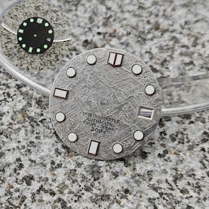 May include: A silver-coloured watch face with a textured surface. It has white circular and rectangular markers, and the text "MARINEMASTER AUTOMATIC PROFESSIONAL 300m". A smaller, black watch face with green markers is also visible.