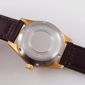 May include: Close-up of a gold-toned wristwatch with a brown leather strap. The watch back is silver with engraved text: "Automatic Movement", "All Stainless Steel", "Sapphire Crystal", and "Water Resistant 200M".