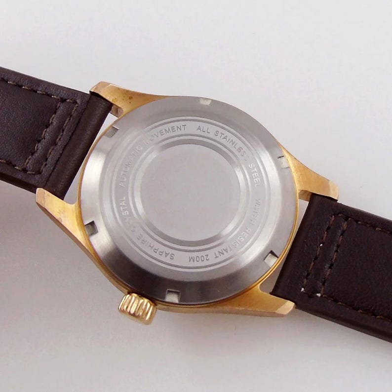 May include: Close-up of a gold-toned wristwatch with a brown leather strap. The watch back is silver with engraved text: "Automatic Movement", "All Stainless Steel", "Sapphire Crystal", and "Water Resistant 200M".
