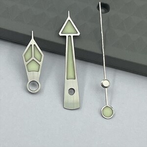 May include: Three silver watch hands with luminous green accents. The hands have various shapes, including a diamond, a triangle, and a thin, straight design. The hands are designed for a watch.