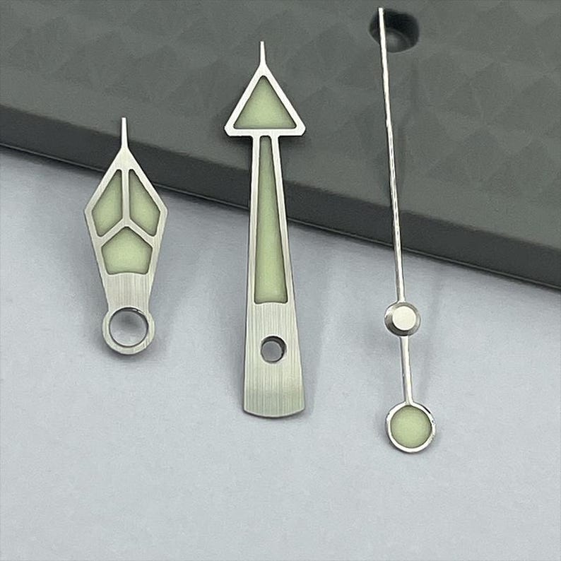 May include: Three silver watch hands with luminous green accents. The hands have various shapes, including a diamond, a triangle, and a thin, straight design. The hands are designed for a watch.