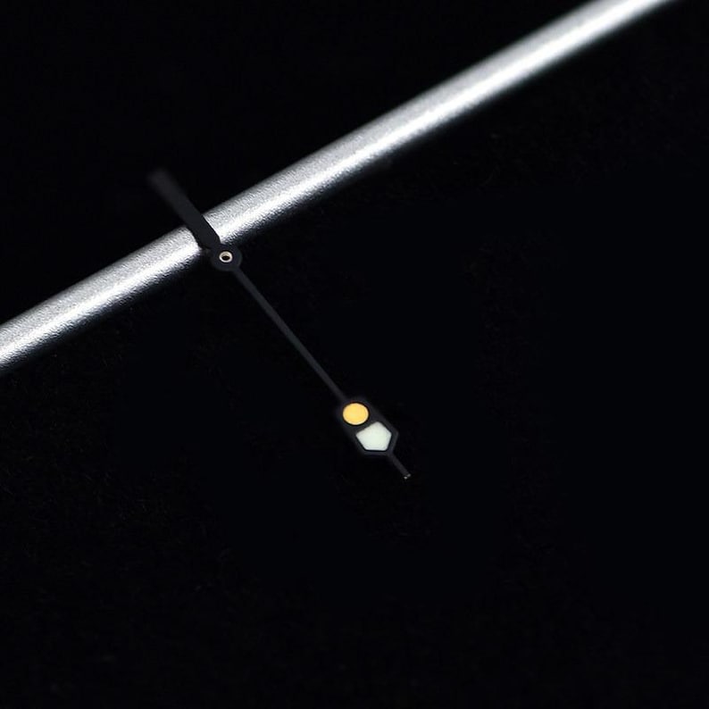 May include: Close-up of a watch face with a black background. The watch hands are black with a yellow and white luminous dot. A silver metal bar is in the upper left corner.