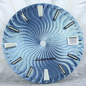 Puede incluir: Una esfera de reloj redonda de color azul con un patrón guilloché en espiral que irradia desde el centro. Incluye marcadores de hora plateados con inserciones luminosas blancas, una ventana de fecha rectangular a las tres y la palabra Automatic.