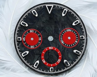 May include: A round watch dial featuring a black forged carbon fiber texture with three red sub-dials. The dial includes silver-toned hour markers, a triangular index at the twelve o'clock position, and white printed numerals on the sub-dials.