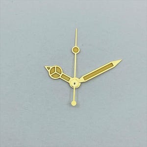 May include: Gold-colored watch hands set against a gray background. The set includes an hour hand with a unique design, a minute hand, and a second hand. The hands are made of a shiny, metallic material.