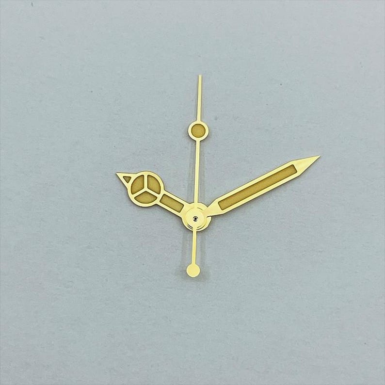 May include: Gold-colored watch hands set against a gray background. The set includes an hour hand with a unique design, a minute hand, and a second hand. The hands are made of a shiny, metallic material.