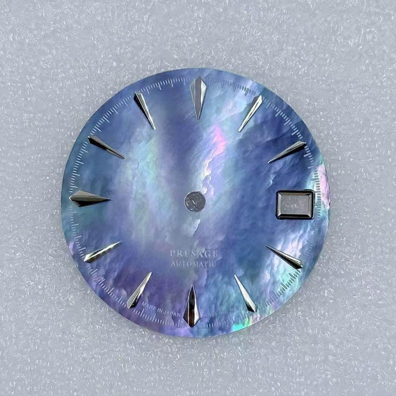 Puede incluir: Una esfera de reloj con una superficie azul iridiscente y brillante. Cuenta con marcadores de hora plateados y puntiagudos y una ventana de fecha. El texto "PRESAGE AUTOMATIC" est&aacute; impreso en la parte inferior. La esfera tiene un di&aacute;metro de unos 3,8 cm.