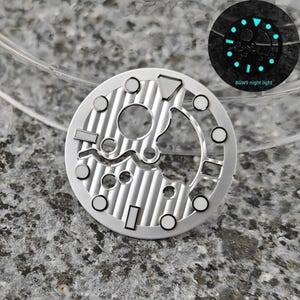 May include: A silver watch dial with a complex, open design featuring various shapes and circular markers. The dial has a textured, striped pattern. A smaller inset shows the dial glowing with blue luminescence, with the text "BGW9 night light".