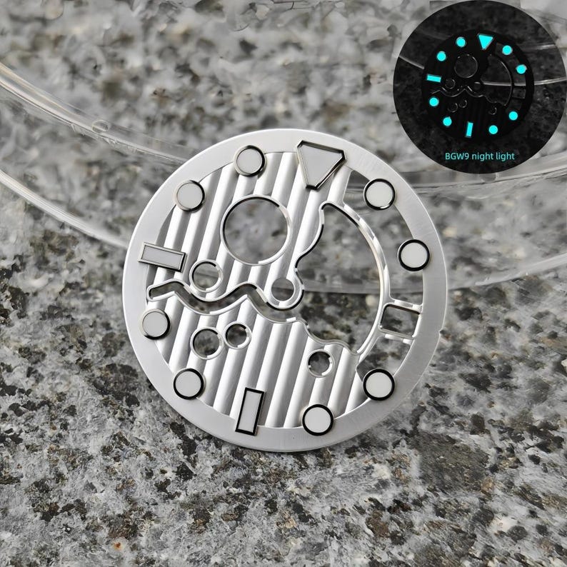 May include: A silver watch dial with a complex, open design featuring various shapes and circular markers. The dial has a textured, striped pattern. A smaller inset shows the dial glowing with blue luminescence, with the text "BGW9 night light".