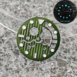 May include: A green watch dial with white and silver accents. The dial features a circular design with various shapes and markings, including a wave-like pattern. The image also shows a glowing night light version of the dial with the text "BGW9 night light".
