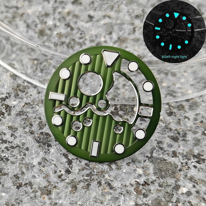 May include: A green watch dial with white and silver accents. The dial features a circular design with various shapes and markings, including a wave-like pattern. The image also shows a glowing night light version of the dial with the text "BGW9 night light".
