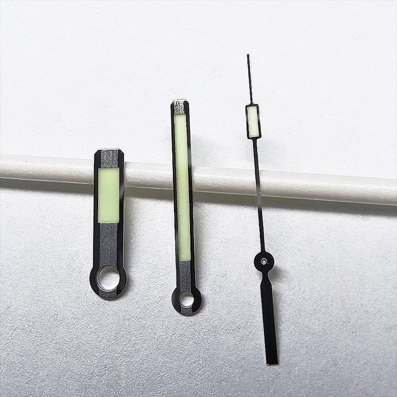 May include: Three watch hands: a short, rectangular hand, a longer, rectangular hand, and a thin, black hand with a circular base. All hands have a silver-colored border and a luminous green center. The hands are for a watch.