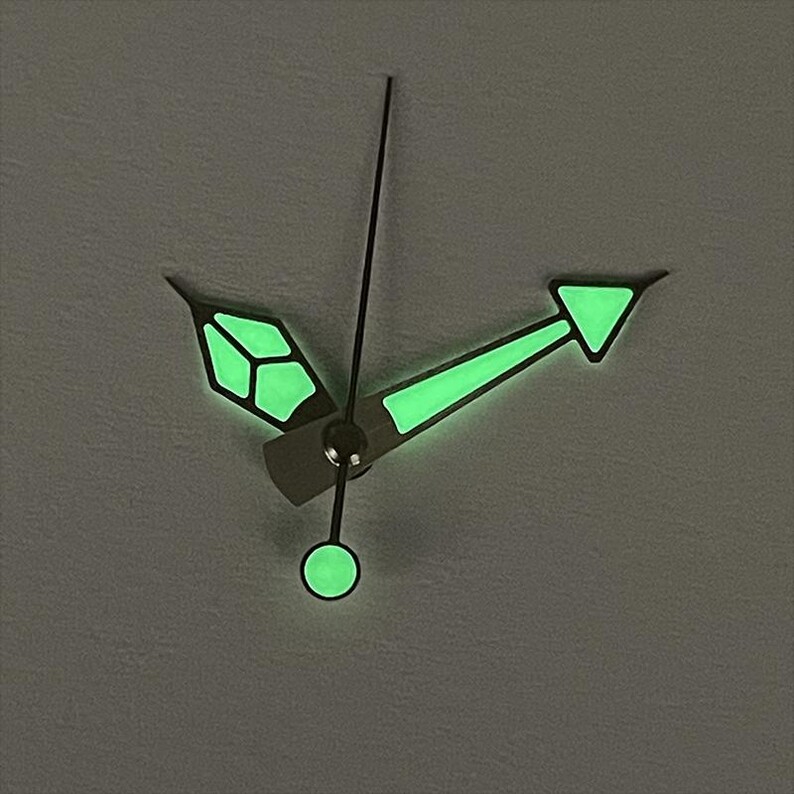 May include: Close-up of watch hands with a luminous green glow. The hour hand has a geometric design, the minute hand is arrow-shaped, and the second hand is a thin line. The hands are set against a gray background.