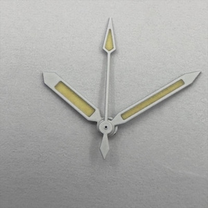 May include: Close-up of watch hands. The hands are silver with a yellow-beige luminous fill. The second hand is a thin, pointed arrow. The minute and hour hands are rectangular with angled tips.