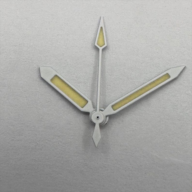 May include: Close-up of watch hands. The hands are silver with a yellow-beige luminous fill. The second hand is a thin, pointed arrow. The minute and hour hands are rectangular with angled tips.