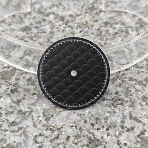 Op de afbeelding: Een zwart, rond horlogegezicht met een waaiervormig patroon en een kleine, heldere edelsteen in het midden. De buitenrand heeft witte markeringen. Diameter ongeveer 3,5 cm.