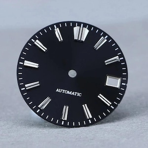 Può includere: Un quadrante per orologio rotondo nero con indici applicati color argento, un marcatore a doppia barra a ore dodici, una finestrella per la data a ore tre e la scritta AUTOMATIC stampata in bianco sotto il foro centrale.