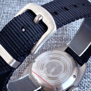 K&ouml;nnte beinhalten: Nahaufnahme einer Armbanduhr mit schwarzem Nylonarmband und silberfarbener Schnalle. Das Zifferblatt ist sichtbar, mit einem silberfarbenen Geh&auml;use und einem strukturierten R&uuml;cken. Die Uhr ist f&uuml;r den t&auml;glichen Gebrauch konzipiert.