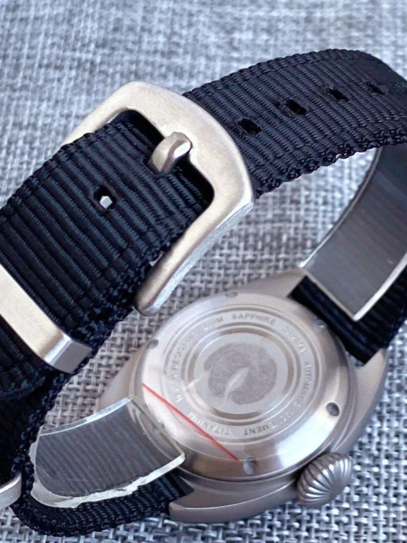 K&ouml;nnte beinhalten: Nahaufnahme einer Armbanduhr mit schwarzem Nylonarmband und silberfarbener Schnalle. Das Zifferblatt ist sichtbar, mit einem silberfarbenen Geh&auml;use und einem strukturierten R&uuml;cken. Die Uhr ist f&uuml;r den t&auml;glichen Gebrauch konzipiert.