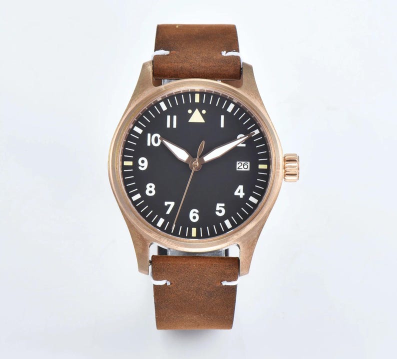 May include: A bronze-colored wristwatch with a black face and white numerals. The watch has a brown leather strap with white stitching. The watch face displays the date and has luminous hands. The watch case is approximately 40mm in diameter.