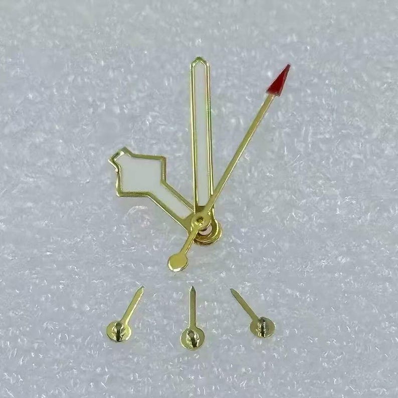 May include: A collection of watch hands and pins. The hands are gold-colored with white and red accents. The hour hand has a white rectangular shape, and the minute hand is a gold arrow with a red tip. Several gold-colored pins are also included.