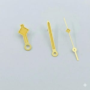 K&ouml;nnte beinhalten: Drei goldfarbene Uhrenzeiger in verschiedenen Designs. Ein Zeiger hat eine rautenf&ouml;rmige Spitze, ein anderer eine spitze Spitze und der dritte eine einzigartige Form. Jeder Zeiger hat ein kleines Loch an der Basis.
