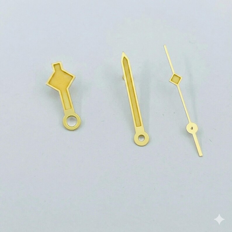 K&ouml;nnte beinhalten: Drei goldfarbene Uhrenzeiger in verschiedenen Designs. Ein Zeiger hat eine rautenf&ouml;rmige Spitze, ein anderer eine spitze Spitze und der dritte eine einzigartige Form. Jeder Zeiger hat ein kleines Loch an der Basis.