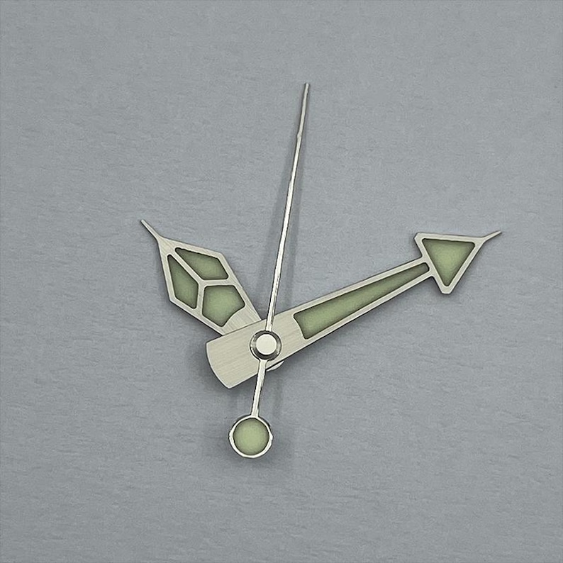 May include: Close-up of watch hands on a gray background. The hands are silver with a luminescent green fill. The hour hand is a stylized arrow, the minute hand is a long, thin pointer, and the second hand has a circular end.