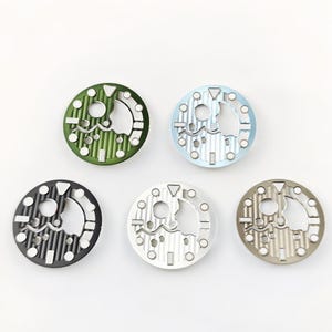 May include: Five circular watch dials in various colors: green, light blue, black, silver, and taupe. Each dial has a unique open design with geometric shapes and a central cutout. The dials are arranged against a white background.