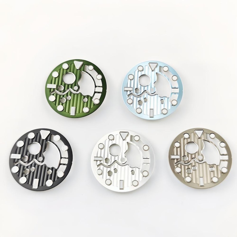 May include: Five circular watch dials in various colors: green, light blue, black, silver, and taupe. Each dial has a unique open design with geometric shapes and a central cutout. The dials are arranged against a white background.