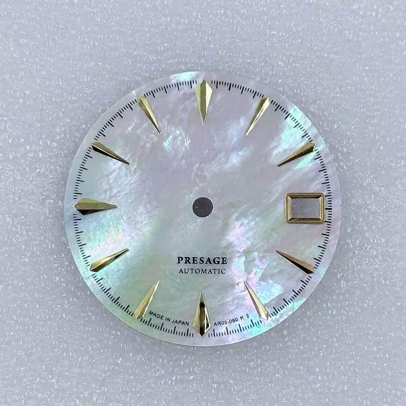 Puede incluir: Una esfera de reloj con una superficie nacarada e iridiscente. Los marcadores de hora y las manecillas triangulares de color dorado est&aacute;n colocados alrededor del per&iacute;metro. El texto "PRESAGE AUTOMATIC" est&aacute; impreso en negro, junto con "MADE IN JAPAN" y "AR05-090 R 2".