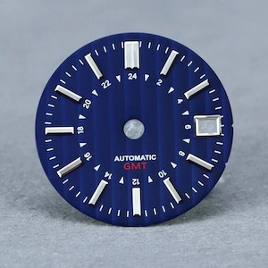 May include: A circular blue watch dial featuring a vertical striped texture, silver applied hour markers, and a date window at the three o'clock position. The dial displays a 24-hour GMT scale with white numerals and the text AUTOMATIC GMT in red.