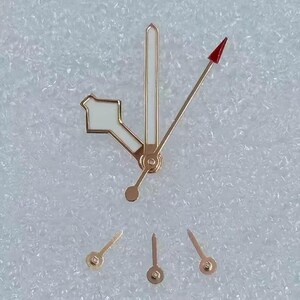 May include: A collection of gold-toned watch hands with white and red accents. The hands are in various shapes and sizes, including a long, thin hand with a red arrow tip. The hands are likely for watch repair or customization.