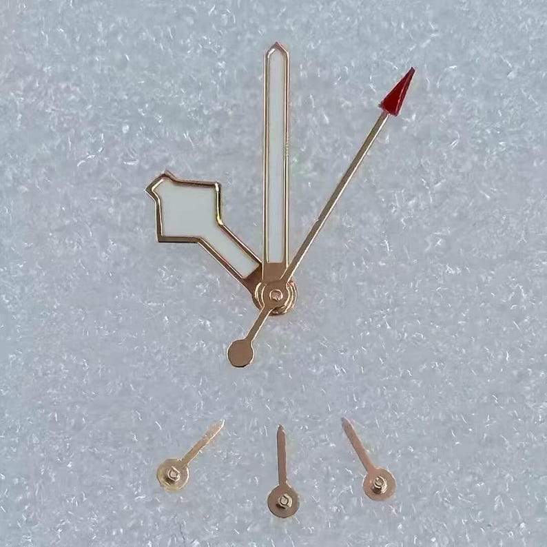 May include: A collection of gold-toned watch hands with white and red accents. The hands are in various shapes and sizes, including a long, thin hand with a red arrow tip. The hands are likely for watch repair or customization.
