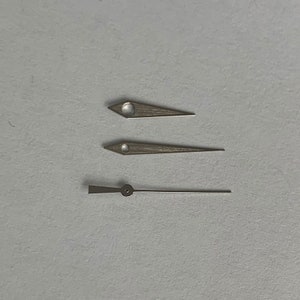 May include: Three watch hands, one long and two short, are arranged on a white surface. The hands are silver in colour and have a pointed shape.