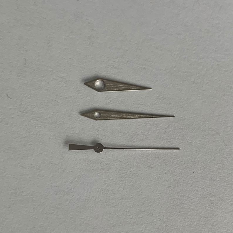 May include: Three watch hands, one long and two short, are arranged on a white surface. The hands are silver in colour and have a pointed shape.