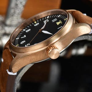 May include: Close-up of a bronze-colored wristwatch with a black face and white numerals. The watch has white hands and a brown leather band with a suede finish. The watch's crown is visible on the side.