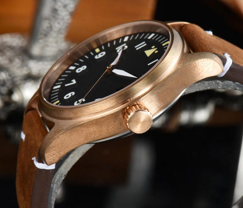 May include: Close-up of a bronze-colored wristwatch with a black face and white numerals. The watch has white hands and a brown leather band with a suede finish. The watch's crown is visible on the side.