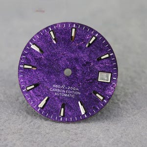Puede incluir: Una esfera de reloj redonda de color morado con acabado de fibra de carbono texturizada, con marcadores de hora plateados, una ventana de fecha en la posición de las tres y el texto 660ft=200m CARBON EDITION AUTOMATIC en blanco.
