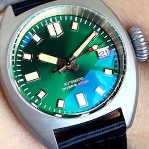 K&ouml;nnte beinhalten: Eine automatische Taucheruhr aus Edelstahl mit gr&uuml;nem Zifferblatt und schwarzem Armband. Das Zifferblatt der Uhr verf&uuml;gt &uuml;ber Leuchtzeiger und -markierungen, ein Datumsfenster und den Text "AUTOMATIC DIVER'S 200m". Die Uhr hat eine strukturierte Krone und ein schwarzes Stoffarmband.