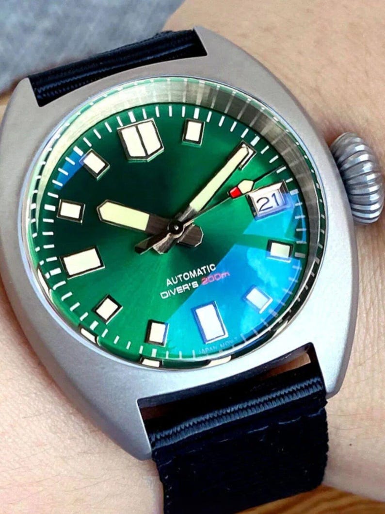 K&ouml;nnte beinhalten: Eine automatische Taucheruhr aus Edelstahl mit gr&uuml;nem Zifferblatt und schwarzem Armband. Das Zifferblatt der Uhr verf&uuml;gt &uuml;ber Leuchtzeiger und -markierungen, ein Datumsfenster und den Text "AUTOMATIC DIVER'S 200m". Die Uhr hat eine strukturierte Krone und ein schwarzes Stoffarmband.
